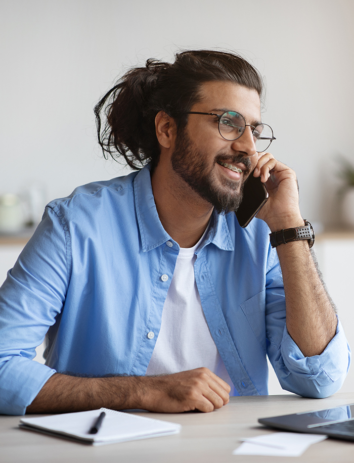 A man with long hair, braces, and glasses smiles as he listens to someone on the other end of his cell phone